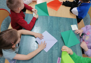 Dzieci siedzą przy stoliku w szkolnej bibliotece. Składają z kolorowego papieru origami.