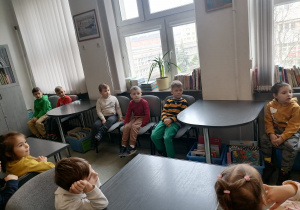Dzieci siedzą przy stolikach w szkolnej bibliotece. Uczestniczą w prowadzonych zajęciach.