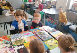 Dzieci oglądają książki w szkolnej bibliotece przy stolikach.