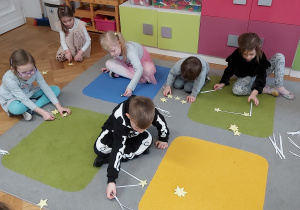 Dzieci siedzą na podłodze i układają konstelacje gwiezdne. wykorzystują do tego celu wycięte z papieru gwiazdki oraz patyczki.