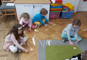 Dzieci siedzą na podłodze i układają konstelacje gwiezdne. wykorzystują do tego celu wycięte z papieru gwiazdki oraz patyczki.