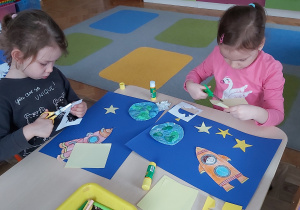 Dwie dziewczynki wykonują pracę plastyczną. Wycinają i naklejają gwiazdki oraz pokolorowaną własnoręcznie rakietę.
