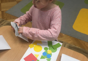 Dziewczynka siedzi przy stoliku i z kolorowych, wyciętych z papieru figur geometrycznych układa rakietę i ufoludka.