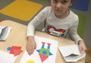 Dziewczynka siedzi przy stoliku i z kolorowych, wyciętych z papieru figur geometrycznych układa rakietę.
