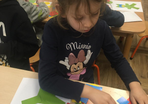 Dziewczynka siedzi przy stoliku i z kolorowych, wyciętych z papieru figur geometrycznych układa ufoludka.
