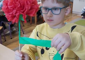 Chłopiec wykonuje kwiatek z bibuły. Owija łodygę zieloną krepiną.