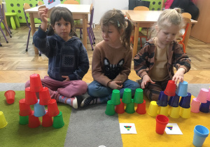 Dwie dziewczynki układają wieżę z kolorowych kubków wg ustalonego kodu. Jedna pokazuje planszę z kodem.