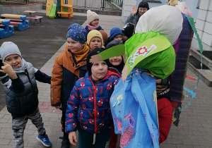 Dzieci stoją w parach przed budynkiem przedszkola. Jedno z nich trzyma w ręku Marzannę.