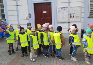 Dzieci ubrane w odblaskowe kamizelki stoją w parach przed budynkiem przedszkola