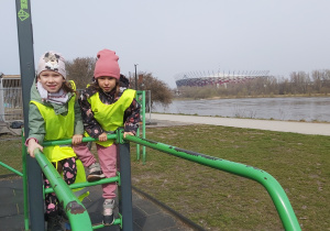 Dwie dziewczynki ćwiczą na poręczach. W tle widać Wisłę i Stadion Narodowy.