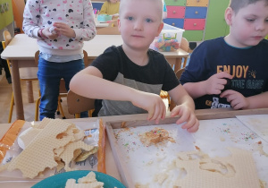 Chłopiec siedzi przy stoliku i posypuje kolorową posypką wafle posmarowane masą kajmakową. Obok stolika stoi dziewczynka i przygląda się jego pracy.