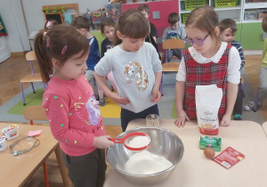 Trzy dziewczynki dodają składniki do ciasta. Jedna z dziewczynek przesiewa mąkę przez sitko.