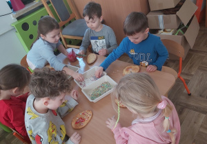 Dzieci siedzą przy stoliku i dekorują upieczone ciastka lukrem i kolorową posypką.