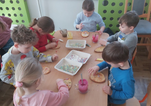 Dzieci siedzą przy stoliku i dekorują upieczone ciastka lukrem i kolorową posypką.