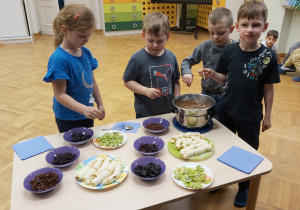 Czworo dzieci z najstarszej grupy stoi koło stolika, na którym znajduje się garnek z rozpuszczoną czekoladą oraz bakalie i owoce na talerzykach.