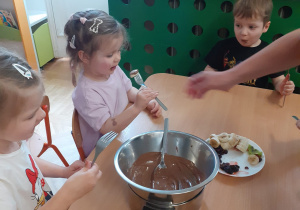 Dzieci z najmłodszej grupy jedzą za pomocą widelców banany umoczone w czekoladzie.