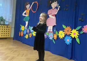 Dziewczynka w czarnej sukience śpiewa. Stoi na tle dekoracji przedstawiającej dzieci oraz napis "Ja też potrafię".