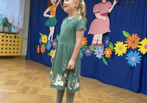 Dziewczynka w aielonej sukience stoi na tle przygotowanej dekoracji i śpiewa piosenkę w jężyku angielskim.