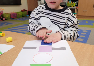 Dziecko wykleja na kartce z figur geometrycznych pajacyka