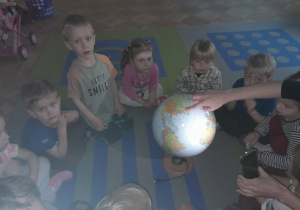Dzieci siedzą na dywanie i patrzą na oświetlany przez nauczyciela globus