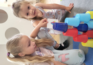 Dwie dziewczynki układają wieżę z kolorowych kubeczków wg wzoru na kartoniku.