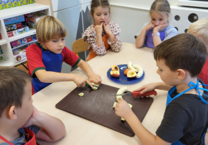 Kilkoro dzieci w kolorowych kuchennych fartuszkach kroi owoce na deskach kuchennych.