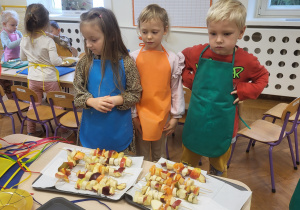 Troje dzieci stoi przy tackach z owocowymi szaszłykami.