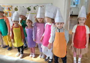 Dzieci stoją w kolorowych fartuszkach i czapkach kucharskich.