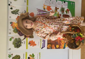 Dziewczynka w stroju Pani Jesieni stoi na tle tablicy, na której widnieją obrazki drzew i liści.