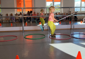 Dziewczynka przeskakuje z jednej obręczy do drugiej trzymając między nogami miotłę.