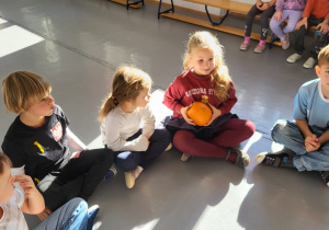 Dzieci siedzą w kole i podają sobie dynie w rytm muzyki.