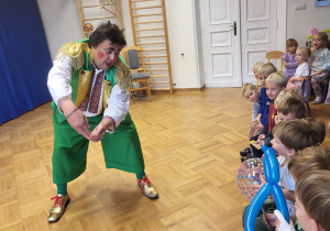 Klaun stoi przed dziećmi i namawia ich do rzutu piłką do celu.