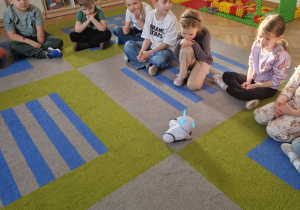 Dzieci siedzą w kole i witają się z robotem.