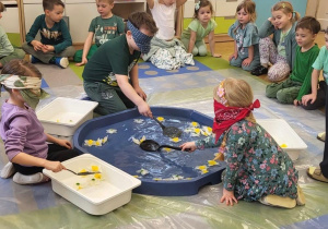 Wiosenne zabawy. Dzieci z zasłoniętymi oczami wyławiają kwiatki z wody za pomocą łyżek cedzakowych.