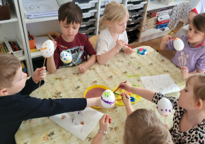 Dzieci siedzą przy stoliku i ozdabiają styropianowe jajka, malując je farbami.