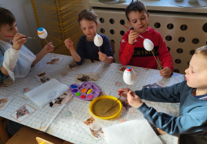 Dzieci siedzą przy stoliku i ozdabiają styropianowe jajka, malując je farbami.