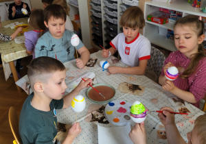 Dzieci siedzą przy stoliku i ozdabiają styropianowe jajka, malując je farbami.