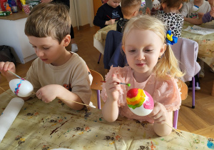 Dzieci siedzą przy stoliku i ozdabiają styropianowe jajka, malując je farbami.
