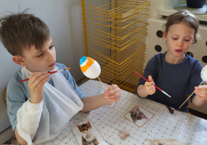 Dzieci siedzą przy stoliku i ozdabiają styropianowe jajka, malując je farbami.