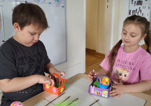 Dwoje dzieci tworzy wielkanocny koszyczek z pisanką.