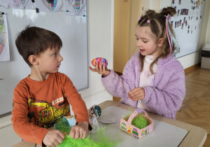 Dwoje dzieci tworzy wielkanocny koszyczek z pisanką.